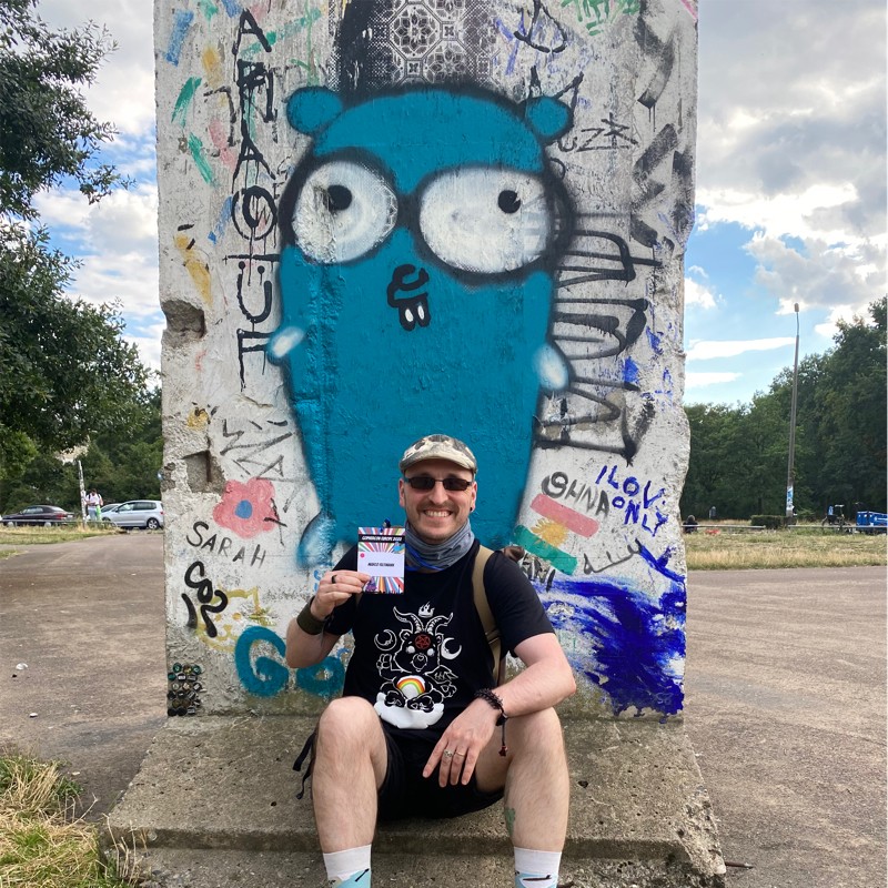 me, at gopher con eu 2023 in front of the German Wall memorial with a graffity gopher. I present my attendee pass looking proud and happy